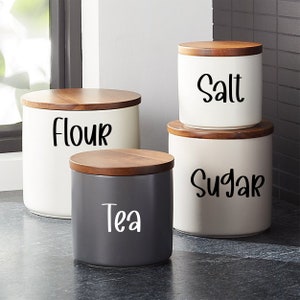 May include: Four white ceramic canisters with wooden lids, labeled "Flour", "Salt", "Sugar", and "Tea".