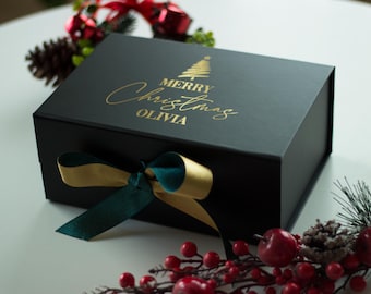 May include: A black gift box with a gold and green ribbon. The box is decorated with a gold Christmas tree and the words "MERRY Christmas OLIVIA". Festive holiday decor surrounds the box.