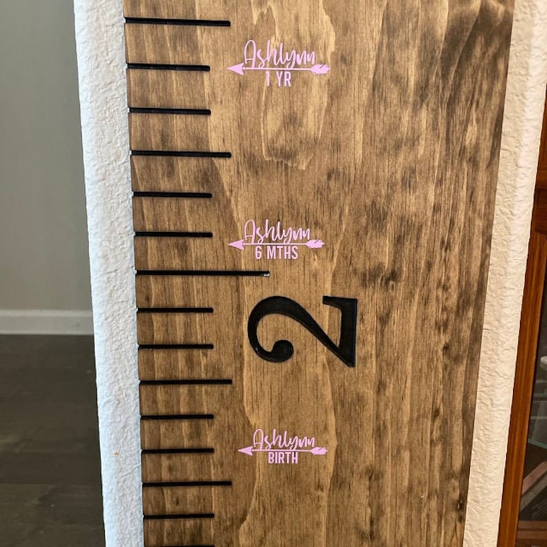 Op de afbeelding: Een houten groeischema met zwarte markeringen en een groot zwart nummer "2" aan de rechterkant. Er zijn drie roze stickers met de naam "Ashlynn" en de data "BIRTH", "6 MTHS" en "1 YR" op het schema.