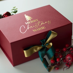 Könnte beinhalten: Eine weinrote Geschenkbox mit goldfarbenem Schriftzug „MERRY Christmas MARIE“ und einer goldenen Weihnachtsbaumgrafik. Ein grünes und goldenes Band ist um die Box gebunden. Dekorative Grünpflanzen und rote Beeren umgeben die Box.