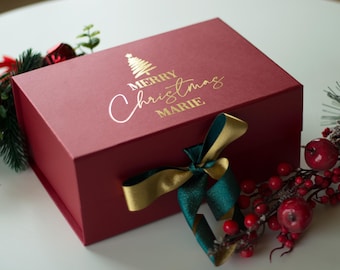May include: A burgundy gift box with gold lettering that reads "MERRY Christmas MARIE" and a gold Christmas tree graphic. A green and gold ribbon is tied around the box. Decorative greenery and red berries surround the box.