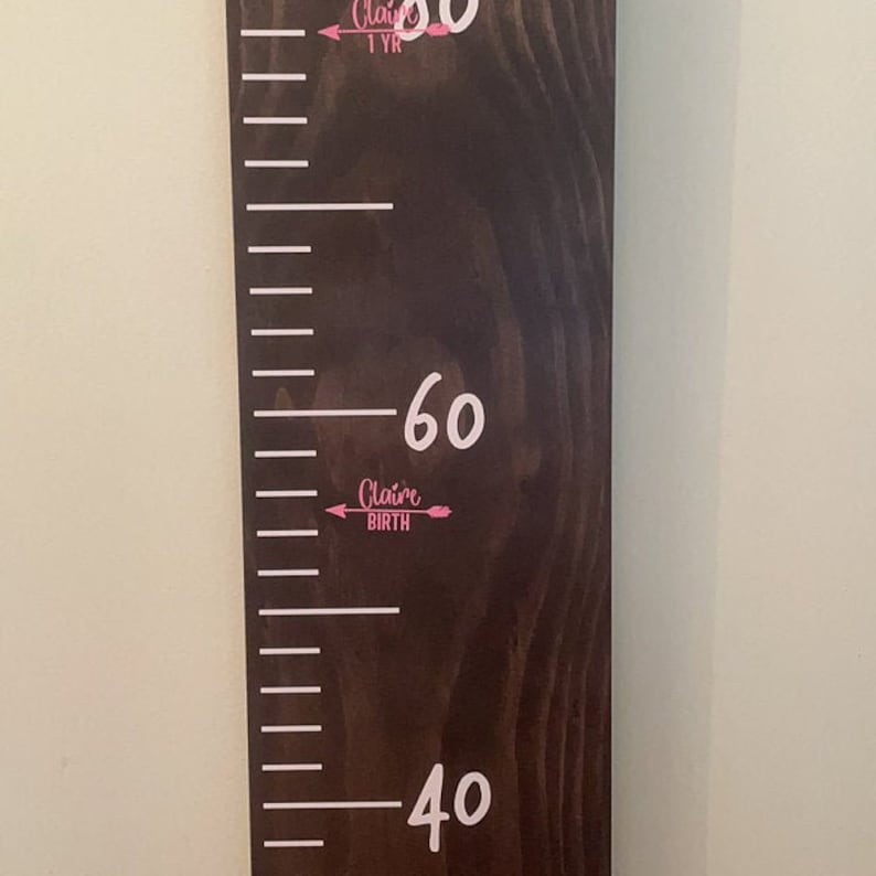 Op de afbeelding: Een bruine houten groeischema met witte markeringen en nummers. De nummers 40, 60 en 80 zijn zichtbaar. De tekst "Claire Birth" is in roze geschreven met een pijl aan elke kant.