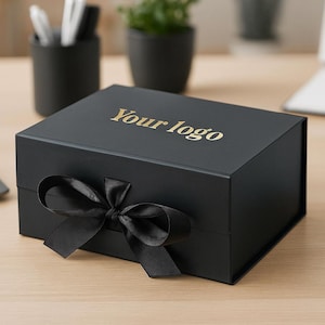 May include: A black gift box with a black ribbon bow. The box has gold text that reads "Your logo". The box is on a light wood surface, with a laptop, pens, and a notebook in the background.