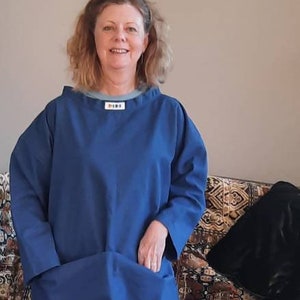 May include: A woman wearing a blue, long-sleeved shirt with a light blue collar and the word "DINE" printed on the front. She is wearing grey trousers and orange socks with white polka dots. She is standing in front of a sofa with patterned cushions.