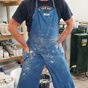 May include: A man wearing a blue apron with the text "K Crew Built" on it. He is standing in a pottery studio with shelves of pottery and a kiln in the background.