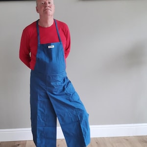 May include: A man wearing a red long-sleeved shirt and a blue apron with a white label that says "BIB." He is wearing black trainers with white accents.