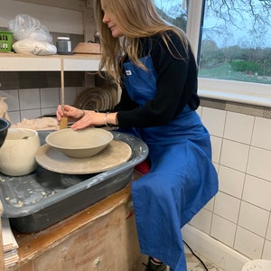 Puede incluir: Una persona que lleva un delantal azul está trabajando en un torno de alfarería, dando forma a un cuenco de arcilla con una herramienta de madera. El torno está sobre un soporte de madera con una bandeja de plástico negro debajo. La persona lleva botas de trabajo negras.