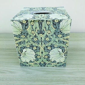 May include: A square tissue box cover with a floral design in shades of green, yellow, and white. The design features stylised flowers and leaves on a dark background.