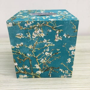 May include: A square tissue box with a teal background, decorated with a print of a flowering tree branch. The branches are brown, and the flowers are white. The top has a circular opening for tissue dispensing.