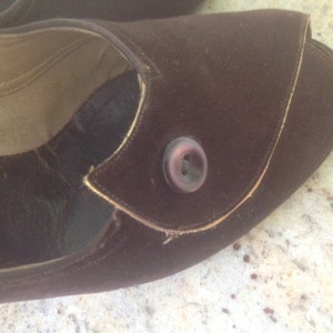 May include: A brown suede shoe with a gold trim and a brown button. The shoe has a pointed toe and a low heel.