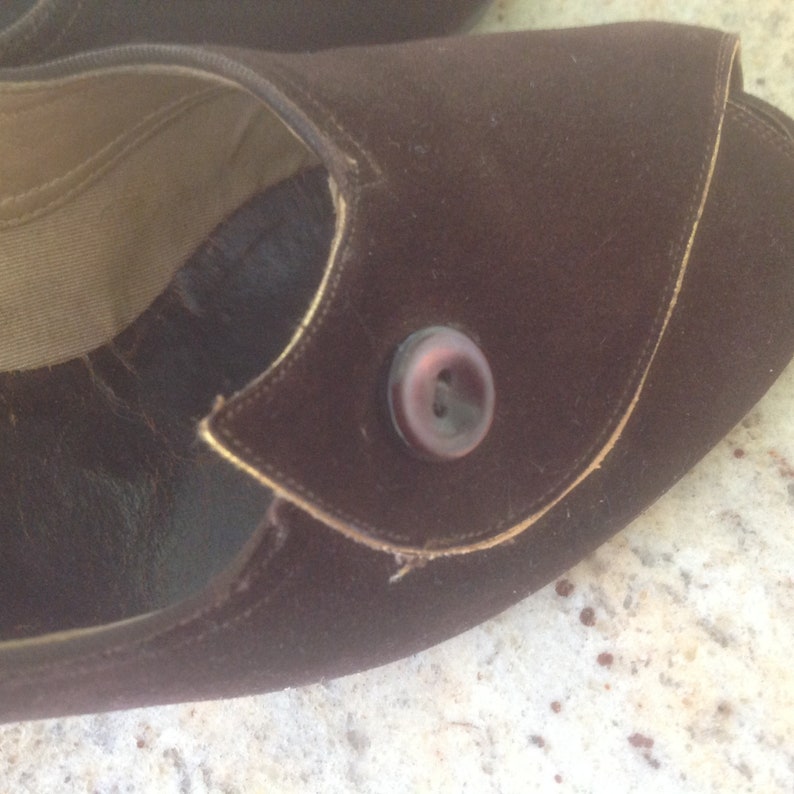 May include: A brown suede shoe with a gold trim and a brown button. The shoe has a pointed toe and a low heel.