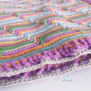May include: A colourful crocheted blanket with a chevron pattern in shades of pink, purple, orange, green, and blue. The blanket has a double border in purple and grey.