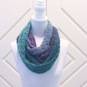 Puede incluir: Una bufanda infinita de ganchillo en tonos de azul verdoso, azul y morado. La bufanda está hecha con un delicado patrón de punto abierto.