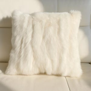 May include: A square, off-white decorative pillow with a fluffy, fur-like texture. The pillow is placed on a white leather couch, with the light creating shadows on the pillow's surface.