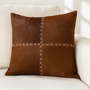 May include: A square decorative pillow with a two-tone brown design. The pillow features a cross-stitched pattern in a lighter brown color, creating a textured look. The pillow is placed on a white sofa.