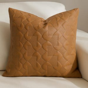 May include: A square, tan-colored decorative pillow with a quilted pattern. The pillow has a geometric design stitched into the surface, creating a textured appearance. The pillow is resting on a white sofa.