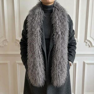 Könnte beinhalten: Ein langer, grauer Schal aus Kunstfell mit lockiger Textur, drapiert über einem dunkelgrauen Wollmantel und einem passenden dunkelgrauen Rollkragenpullover. Der Schal hat einen dichten, zotteligen Flor, der vertikal vor dem Mantel herabhängt.