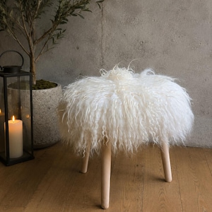 May include: A round stool featuring a thick, white, long-haired sheepskin seat cushion supported by three light-colored wooden legs. The fluffy, textured wool fibers drape over the edges of the circular seat, creating a soft and voluminous appearance.