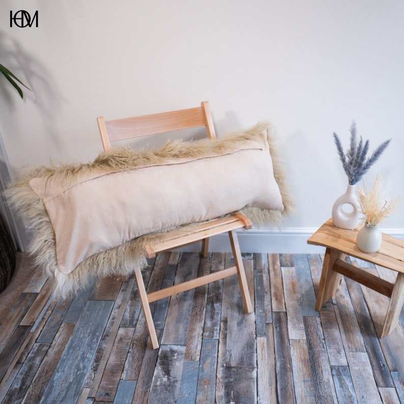 May include: A long, beige cushion with a fluffy, light brown faux fur trim sits on a wooden folding chair. The chair is on a wood-effect floor.