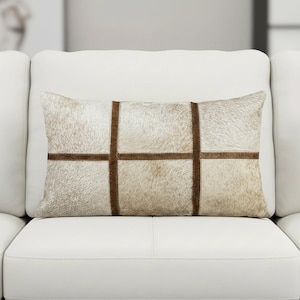 May include: Rectangular decorative pillow with a natural hide appearance. The pillow features a light beige and brown color scheme with a grid pattern created by dark brown stitched lines. The pillow is placed on a white sofa.