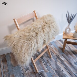 May include: A light beige fluffy sheepskin throw pillow resting on a wooden folding chair. The chair is in front of a white wall and a wooden table with a vase of dried flowers.