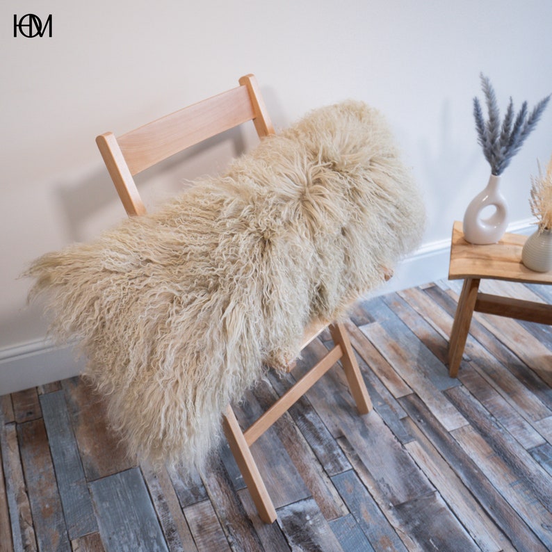 May include: A light beige fluffy sheepskin throw pillow resting on a wooden folding chair. The chair is in front of a white wall and a wooden table with a vase of dried flowers.