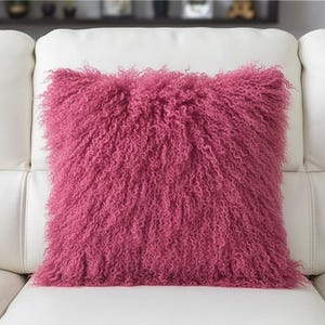 May include: A magenta-coloured cushion with a shaggy, curly texture. The square cushion is placed on a white leather sofa. The texture of the cushion gives it a soft, inviting appearance, suitable for home decor.