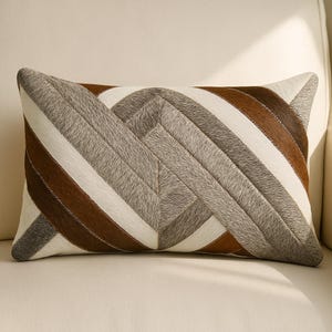 Könnte beinhalten: Rechteckiges Zierkissen mit geometrischem Muster. Das Kissen hat ein Design aus abwechselnden braunen, grauen und weißen Streifen. Das Kissen ist aus einem strukturierten Material, wahrscheinlich Kuhfell, gefertigt und liegt auf einer cremefarbenen Oberfläche.