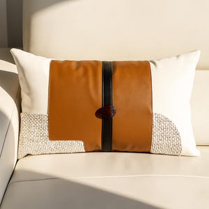 May include: Rectangular decorative cushion featuring a brown leather-effect central panel, a black vertical stripe, and a dark brown button closure. The cushion has white and textured beige fabric sections. The cushion is on a white sofa.