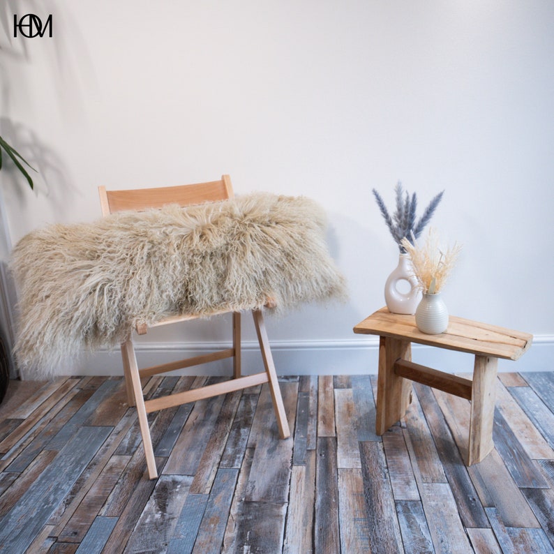 May include: A light brown fluffy sheepskin throw pillow on a wooden folding chair. The chair is on a wood floor with a light blue and brown pattern. A small wooden stool with a white vase and dried flowers sits in the background.