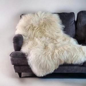 Può includere: Un tappeto in pelle di pecora color crema, soffice, drappeggiato su un divano grigio scuro. Il tappeto ha lunghe fibre morbide e aggiunge un tocco di calore e consistenza alla stanza. Il divano ha un design classico e un aspetto invitante.