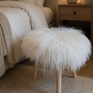 Könnte beinhalten: Ein runder, weißer, flauschiger Hocker mit hellen Holzbeinen. Der Hocker steht auf einem neutralfarbenen Teppich in einem Schlafzimmer. Die zottelige Textur des Hockers kontrastiert mit den glatten Holzbeinen und den umliegenden weichen Möbeln.