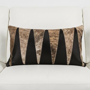 Könnte beinhalten: Rechteckiges Zierkissen mit einem Design aus Kuhfell und Leder. Das Kissen hat abwechselnd schwarze Leder- und braune Kuhfell-Dreiecke. Das Kissen liegt auf einem weißen Sofa.