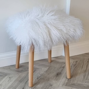 Può includere: Un piccolo sgabello rotondo in finta pelliccia bianca con quattro gambe in legno chiaro. La seduta dello sgabello è ricoperta da una lunga e folta pelliccia bianca. Le gambe sono distanziate in modo uniforme e leggermente angolate verso l'esterno. È un elemento d'arredo decorativo.