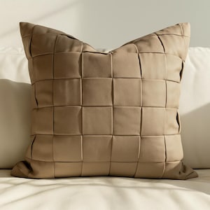 May include: A square, tan-colored decorative pillow with a woven, grid-like pattern. The pillow is made of a soft material and is placed on a white surface. The pillow has a classic, neutral design.