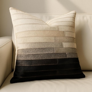 May include: A decorative square pillow with a gradient design. The pillow transitions from light beige at the top to black at the bottom, with shades of gray and brown in between. The pillow is made of rectangular pieces of material.