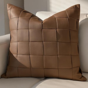 May include: A square, brown leather cushion with a woven design. The cushion is made of individual squares of leather stitched together, creating a textured surface. The cushion is resting on a white sofa.