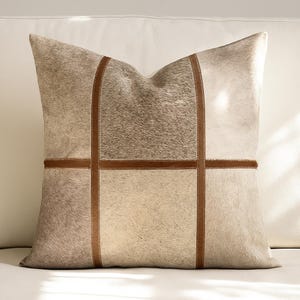 Könnte beinhalten: Ein quadratisches Zierkissen mit einem Patchwork-Design. Das Kissen hat Abschnitte aus hellbraunem und grauem Kunstfell, die durch braune, lederartige Streifen getrennt sind. Das Kissen liegt auf einer weißen Oberfläche.