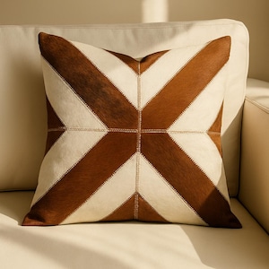 May include: Square cushion with a geometric pattern. The cushion has a patchwork design with alternating brown and cream-coloured sections. The sections are stitched together with cream-coloured thread. The cushion is resting on a cream-coloured sofa.