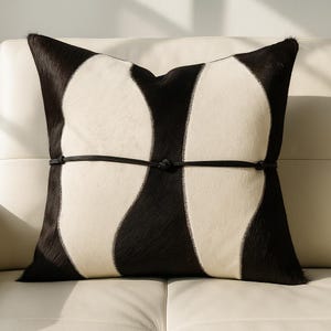 May include: Square decorative pillow with a cowhide design. The pillow features a black and white pattern with a black leather tie detail. The pillow is a square shape and is designed for home decor.