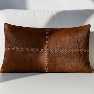 May include: A rectangular brown cowhide cushion with a stitched cross pattern. The cushion has a soft, textured surface and natural colour variations. The stitching is a lighter brown, creating a contrasting design. Ideal for adding a rustic touch to any room.
