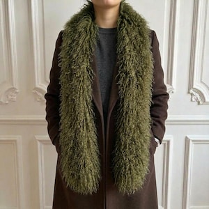Könnte beinhalten: Ein langer, olivgrüner Kunstfellschal mit zotteliger, lockiger Textur, der über einem dunkelbraunen Mantel und einem anthrazitfarbenen Pullover getragen wird. Der Schal hat einen dichten, voluminösen Flor, der vertikal über der dunklen Oberbekleidung hängt.
