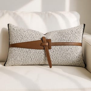 Könnte beinhalten: Rechteckiges Zierkissen mit einem strukturierten schwarz-weißen Stoff. Ein brauner Lederriemen umschließt die Mitte und wird mit einer Schnalle befestigt. Das Kissen hat einen schwarzen Rand und liegt auf einem weißen Sofa.