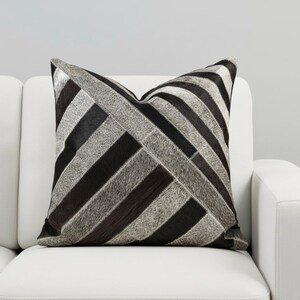 May include: Square decorative pillow with a geometric pattern. The pillow features a patchwork design of black, gray, and white sections. The pillow is placed on a white sofa, creating a contrast of colors and textures.