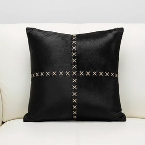 May include: Black square decorative pillow with a cross-stitch design. The pillow has a textured surface and a neutral-colored stitch pattern. The pillow is placed on a white surface.