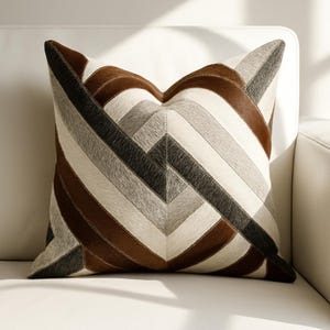 Könnte beinhalten: Quadratisches Zierkissen mit geometrischem Muster. Das Kissen hat ein Patchwork-Design mit braunen, grauen und cremefarbenen Abschnitten. Das Kissen liegt auf einer weißen Oberfläche, wobei das Licht Schatten erzeugt.