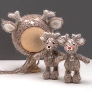 May include: A knitted reindeer set. The set includes a brown hat with antlers and two small reindeer figures. The hat has white spots and cream-colored accents. The reindeer figures are brown with white spots, small black eyes, and one with a red nose.