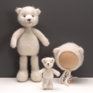 Peut inclure: Un grand ours en peluche tricoté couleur crème, un petit ours en peluche tricoté couleur crème et un bonnet tricoté couleur crème avec des oreilles d'ours. Les ours ont un nez et une bouche brodés marron, le bonnet est sur un support en bois.