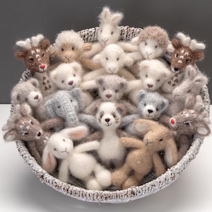 Könnte beinhalten: Ein geflochtener Korb voller handgestrickter Tierfiguren. Die Sammlung umfasst Bären, Kaninchen, Rehe, Schafe und einen Elefanten in Weiß-, Grau- und Brauntönen. Die Spielzeuge haben gestickte Gesichter und sind aus Wolle gefertigt.