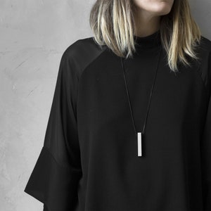 Peut inclure: Une femme portant un chemisier noir avec un collier pendentif en forme de barre argentée.